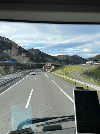 The image displays a highway scene viewed forward from within a vehicle, likely a truck or bus, through its windshield. The upper windshield frame, dashboard, and a portion of the wipers are visible, along with a vertically mounted white tablet or large smartphone on the lower right dashboard. The highway, situated in the hilly region near Zumaia, Spain, consists of two lanes in the direction of travel, separated by a dashed white line, and bordered by solid white lines and metal guardrails on both sides. A white hatchback car travels in the immediate lane ahead, while a smaller white vehicle is further ahead in the left lane. An overpass bridge crosses above the highway, supporting a blue and red semi-trailer truck. Below the overpass, on the right side of the highway, a red and white sign is visible, bearing the text "EUSKAL HERRIA" (Basque Country). The surrounding landscape features undulating hills covered with a combination of evergreen and deciduous trees, and large expanses of green grass. A winding path is visible ascending a grassy slope to the right of the highway. The sky above is partly cloudy, displaying patches of blue amidst white clouds.