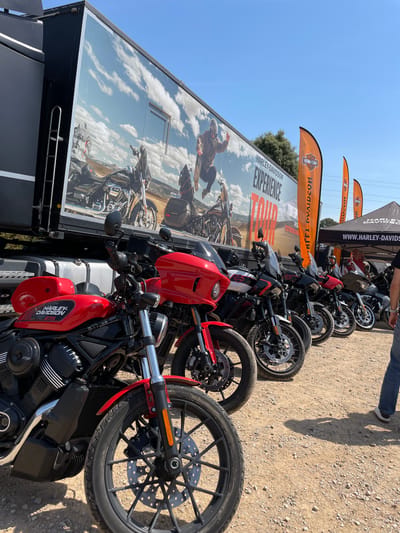The image depicts a Harley-Davidson promotional event in l'Ametlla del Vallès, Spain, as indicated by the truck's branding and the presence of Harley-Davidson motorcycles and flags. A row of Harley-Davidson motorcycles, mostly red and black, are lined up on a gravel surface. Behind the motorcycles, a large trailer displays the "Harley-Davidson Experience Tour" advertisement. Orange Harley-Davidson flags are visible in the background, along with a tent also bearing the Harley-Davidson logo. A person's leg is seen in the upper right corner of the image. The weather appears sunny.