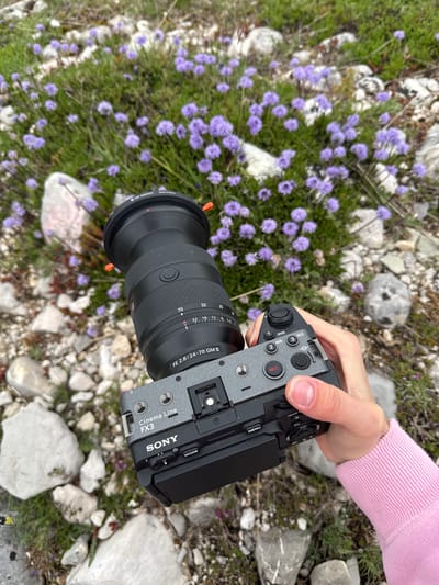 A person holds a Sony FX3 camera with an FE 2.8/24-70 GM II lens over a patch of small purple flowers mixed with light-colored stones and green vegetation. The camera is angled downwards, showing the controls and lens details. The person's hand and a pink sweatshirt sleeve are visible. The background consists of more stones and vegetation, suggesting an outdoor location, potentially in Nikšić, Montenegro.