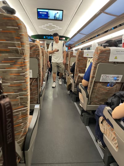 This image captures the interior of a train, likely a high-speed rail service in China, given the "Longhua District, China" context. The perspective is from a passenger's seat, looking down the aisle. The primary subjects are the seats themselves, upholstered in a brown fabric with vertical stripes of white and orange. A man in a white t-shirt and grey shorts stands in the aisle, holding a smartphone, and looking down at it. He is wearing white sneakers. Other passengers are visible in the background, seated in the train cars. One passenger is seen with their legs extended, and another has their arm resting on the back of a seat.
A prominent object in the scene is a screen mounted on the wall of the train, displaying "NEXT STATION HuMen". There are also safety instructions printed on the back of the seats, one of which reads, "调整椅背时,谨防傾倒 when adjusting the back be careful not to tip over the tea". This suggests the train is in motion, and passengers are being reminded of safety procedures. The lighting appears to be artificial, consistent with an indoor environment like a train car. The overall scene suggests a typical journey on a public transportation system, with people engaged in their own activities while traveling. The presence of the man with his phone and the overhead lights imply it could be daytime, though no external weather cues are visible. The text on the screen and the safety instructions provide specific details about the location and operational aspects of the train.
