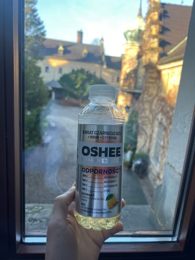 A fair-skinned human hand is positioned centrally, holding a clear plastic bottle of OSHEE Vitamin Water with a white screw cap, in front of a window. The bottle contains a translucent yellow liquid. Its silver-colored label features Polish text, including "KWIAT CZARNEGO BZU + IMBIR + CYTRYNA" (Elderflower + Ginger + Lemon) and "ODPORNOŚĆ" (Immunity), listing 100% of daily values for Vitamin D, Vitamin C, Vitamin B6, and Zinc, along with an illustration of a lemon and ginger. The label also specifies "BEZ: SZTUCZNYCH AROMATÓW, BARWNIKÓW, KONSERWANTÓW" (Without artificial flavors, colors, preservatives).
The window's dark brown frame is visible on the left, bottom, and partially the right side. Through the window, an outdoor scene is observed under a clear blue sky. In the background, there are architectural structures characteristic of a historic complex. A large, light-colored building with a dark roof, multiple windows, and distinctive dormer windows (some with golden accents) stands in the mid-ground on the left. To its right, a prominent stone-built tower or section of a building is heavily covered in autumnal ivy, illuminated brightly by direct sunlight. A paved area or courtyard with some evergreen trees is visible below the buildings. The architectural features confirm the location as Kliczków Castle in Kliczków, Poland. The scene is well-lit by natural sunlight, casting bright highlights on the right-side structures.