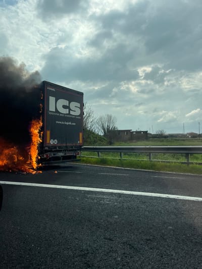 A commercial semi-trailer, branded "KOGEL" and "ICS LOGISTIK UND TRANSPORT GMBH," is stationary on the shoulder of an asphalt multi-lane roadway in Pastorano, Italy. The rear left side of the trailer is engulfed in active flames, emitting dense black smoke that obscures the upper portion of that section. The fire is concentrated at the lower rear of the trailer, with burning debris visible on the road surface beneath it. The trailer's license plate, partially visible, appears to be Italian (e.g., "BG EL F1 11"). A silver metallic guardrail runs parallel to the roadway beyond the shoulder, separating it from an area of green grass and sparse trees. In the distant background, a ruined or unfinished building and power lines are visible under an overcast sky with patches of brighter light. No individuals or other vehicles are explicitly depicted interacting with the scene.