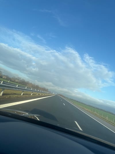 The scene depicts a view from a vehicle traveling on a multi-lane asphalt highway in Vodenichane, Bulgaria, under a partially cloudy sky. The road, appearing dark and possibly wet, extends into the distance, marked by solid and dashed white lines. To the left, a metal guardrail runs parallel to the highway, behind which dormant deciduous trees and green-brown vegetation are visible. To the right, a grassy verge borders an expansive green agricultural field, delineated by a fence with white posts. The sky above features clear blue areas interspersed with large, developing cumulus and stratocumulus clouds. The lower foreground shows the dark dashboard and windshield base of the originating vehicle, with reflections of the road markings. No individuals are visible.