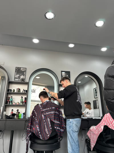 The scene depicts the interior of a modern barbershop in Kumanovo, North Macedonia.

A male barber, with short dark hair, wearing a black long-sleeved sweatshirt with white graphics and light blue jeans, stands at the center. He has tattoos on his left forearm and wears a watch on his left wrist. He is actively cutting the hair of a male client seated in a black barber chair, positioned with his back to the camera. The client is draped in a barber cape featuring red, white, and blue stripes, "BARBERSHOP" text, stars, and barber pole motifs. The barber is using electric clippers on the client's hair.

To the right, partially visible, is a figure wearing a black puffer jacket. In the reflection of an arched, black-framed mirror on the right side of the image, another male client with short dark hair is visible, seated in a barber chair and wearing a white t-shirt with "THE NORTH FACE" logo.

The barbershop features light gray/white walls and a matching ceiling with multiple circular recessed LED lights providing illumination. Black arched mirrors are prominent on the walls. A black shelving unit to the left of the main barber station holds various hair care products in bottles, jars, and spray containers, alongside two green skull-shaped decorative items. A framed black and white poster depicting a cat wearing sunglasses hangs above this shelving unit. A black counter runs beneath the mirrors, with additional products and black electrical cables visible. A window is reflected in the left mirror, showing blurred exterior details.
