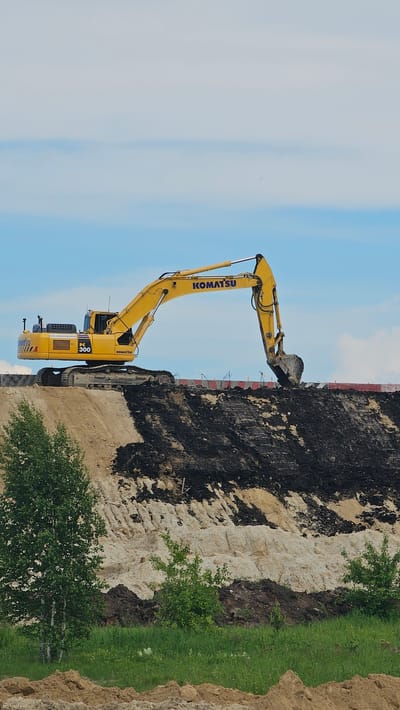 An outdoor, eye-level, vertical, full shot shows a Komatsu PC 300 excavator working on a construction site. The excavator is bright yellow with a black bucket and is positioned on a large pile of dirt. It is actively digging into the pile, with its arm extended. The background consists of a light blue sky with scattered white clouds. Below the excavator, the dirt pile displays layers of different colored soil, including light brown, tan, and dark black. In the foreground, there is green grass and a few small trees. The scene appears to be a construction or earth-moving project. The location is identified as Loginovo, Russia.