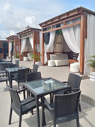 The image shows an outdoor patio with several wooden structures, resembling pavilions, covered in white sheer curtains. There are four black wicker chairs and two glass-topped tables arranged on a concrete patio, with a white couch visible within one of the pavilions. The setting is outdoors, with the presence of green plants in pots. The location is likely in Ujevwu, Nigeria based on the coordinates (latitude: 5.471, longitude: 5.817), which correspond to a residential area near a park. The scene is typical of a restaurant or lounge setting.