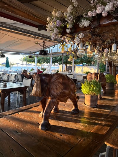 The image depicts a wooden table at an outdoor restaurant or cafe in Angelochori, Greece. The table is adorned with a wooden sculpture of an elephant in the foreground, and in the background, there are potted plants and other decorative elements. The restaurant appears to be situated near a beach, with umbrellas and lounge chairs visible on the sand, and the sea in the distance. The lighting suggests it is either early morning or late afternoon, with warm sunlight casting long shadows across the wooden surfaces. Several tables and chairs are set up, indicating a dining area. Two individuals are seated at a table in the background, with one facing away from the camera and the other partially visible. The overall scene conveys a relaxed, possibly seaside dining atmosphere.