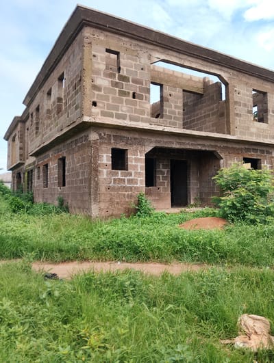This is a medium-angle, eye-level, full shot of a two-story, unfinished concrete block building situated in Orudu-Olokan, Nigeria. The building has an exposed concrete frame and walls constructed from grey concrete blocks. Several openings for windows and doorways are visible, but the structure lacks doors and windows. The roof is incomplete, with exposed beams and openings in the upper level. The building is surrounded by overgrown green grass and vegetation. A dirt path runs in front of the building. To the right of the building, there is a dense bush. The sky above is partly cloudy with a mix of blue sky and white clouds. A pile of dirt is visible near the entrance of the building. In the foreground, to the right, there is a crumpled brown sack.
