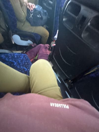 The image captures an interior view, likely within a mode of transportation, showing the legs and torsos of individuals seated. The foreground is dominated by the legs of at least two people, one wearing light olive-green pants and another in a rose-colored garment with the white text "PULL&BEAR" visible. Behind them, other passengers and their belongings are partially visible, including a blue patterned fabric, a purple bag, and a dark blue backpack. The environment appears to be cramped, with seats and dark-colored interior panels suggesting a bus or train. The lighting is dim, contributing to a slightly blurry and intimate atmosphere. The image does not provide any specific visual cues to pinpoint its location to Merhavim Regional Council, Israel.