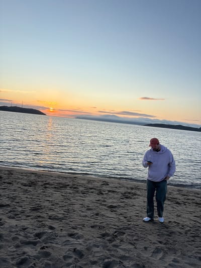 A lone man, wearing a red baseball cap, a light purple hoodie, and dark pants, stands on a sandy beach, looking down at his phone. The ocean stretches out behind him, with gentle waves lapping at the shore. In the distance, a body of land with hills and structures is visible, silhouetted against a sunset sky. The sky is a gradient of soft blues and oranges, with wispy clouds scattered across the horizon. The scene appears to be a tranquil evening on the coast, possibly in the city of Teriberka, Russia, given the characteristic Arctic landscape and the quality of light during a sunset.