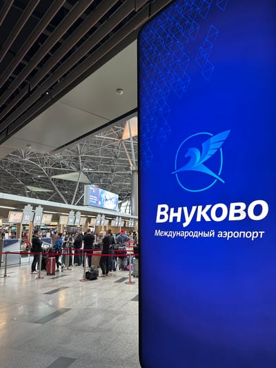 The image depicts a scene inside an airport, likely Vnukovo International Airport in Russia, given the prominent signage. The setting is a check-in area with several rows of airline counters visible, numbered from 92 to 97 and beyond. People are queuing up at these counters, with luggage in tow. The architecture of the airport is characterized by a high, exposed metal ceiling structure with complex bracing, indicative of a large, modern terminal.

The overhead signage is a large, illuminated blue banner featuring the airport's logo and name in Russian: "ВНУКОВО" (Vnukovo) in large white Cyrillic letters, with "Международный аэропорт" (International Airport) written vertically beside it. The logo itself is a stylized bird in flight within a circle, rendered in light blue. Another smaller advertisement banner is visible further down, displaying an airplane and text in a foreign language, possibly related to a specific airline or service.

The lighting suggests it is daytime, with bright artificial lights illuminating the interior. The overall atmosphere appears to be one of typical airport busyness, with individuals and groups of travelers preparing for their flights. The mood is neutral, focused on the functional aspects of air travel. The floor is a polished, light-colored stone or tile. The presence of red velvet ropes indicates a structured queuing system for passengers. The image is taken from a slightly elevated perspective, looking down on the check-in counters and queues.