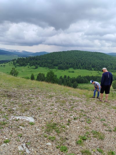 Two individuals, likely a father and son, stand on a gravelly incline overlooking a lush green valley in Туим, Russia. The man on the right wears a navy blue long-sleeved shirt with the word "RUSSIA" in white lettering across the chest, black shorts, and sandals. He is facing the valley. To his left, a younger individual wearing a striped blue and white t-shirt, blue jeans, a blue and white baseball cap, and sandals is bent over, looking at the ground. The landscape is dominated by a rolling green hill covered in dense forest, with a lighter green field in the foreground of the valley. The sky is overcast with heavy, gray clouds. The foreground is a mix of gravel, small stones, and sparse green grass and weeds. The scene appears to be a natural landscape, likely a viewpoint or hiking area.