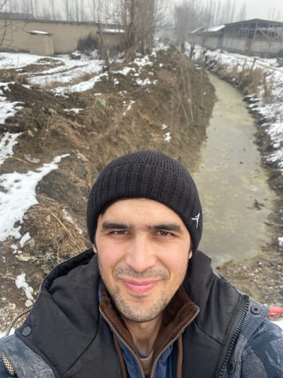 A male individual is depicted in a close-up selfie, smiling directly at the camera. He wears a black knit beanie with a white emblem on the left side, a dark blue or black sleeveless puffer vest over a blue garment, and a brown fur-lined collar. His face shows stubble or a short beard.

The background reveals a winter outdoor scene in what appears to be a rural or semi-rural area of Davlak, Uzbekistan. Snow covers patches of dark earth and dry vegetation. To the left, a large dirt mound, partially snow-covered, is visible. Behind it, a light-colored wall and a small, squat structure stand in the distance. To the right, a narrow, unlined channel or ditch with murky, greenish-brown water extends into the background. Its banks are dirt, partially snow-covered, with sparse vegetation. Further along the channel's right side, structures resembling barns or agricultural buildings with white walls and dark roofs are present. Bare trees are scattered throughout the distant background under an overcast sky.