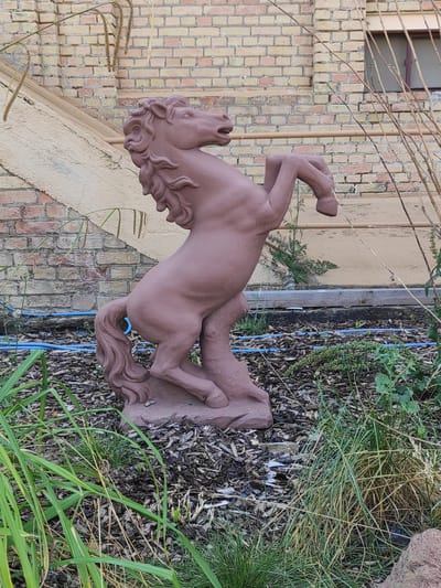 A reddish-brown, rearing horse statue is situated in a garden setting against a brick building. The statue is positioned on a small, uneven mound covered in mulch and wood chips. The horse is depicted with its front legs raised and its mouth open, as if in motion. Lush green grass and dry vegetation surround the base of the statue. In the background, a light-colored brick wall with horizontal accents and a section of a window can be seen. There is also a beige, angled structure and some dry, spiky plants. A blue hose is visible on the ground near the statue. The scene appears to be outdoors, likely in a residential or public garden area in Brühl, Germany.
