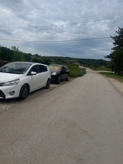 The image shows a paved road in the rural area of Topchii, Bulgaria. Two cars are parked on the side of the road: a white car and a dark-colored car. The sky is overcast with cloudy skies. Trees and vegetation line the road and the surrounding landscape. Overhead power lines are visible.