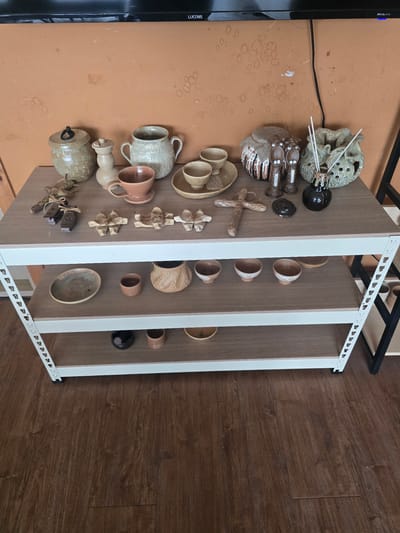 This media file captures a collection of ceramic and wooden decorative objects, along with some small potted plants and what appear to be incense sticks, arranged on a shelf unit against a warm-toned wall and a wooden floor. The arrangement suggests a display or storage of handmade crafts.

The subjects are primarily various ceramic items, including mugs, bowls, and decorative pieces with intricate textures and earth-toned glazes. There are also a few wooden carvings, one resembling a crab and another a cross. Small green plants in simple pots and white sticks, possibly incense, are also visible.

The scene is indoors, with the objects displayed on a white-framed shelf unit with multiple compartments. The wall behind the shelf appears to be a terracotta or warm orange color, with some circular water stains or marks. The floor is made of dark brown wood planks.

No people are visible, and the objects are stationary, indicating a still life composition rather than an active event. The lighting appears to be natural and diffuse, suggesting it could be daytime. There are no strong indicators of weather or specific time of day beyond that. The overall impression is one of an artful arrangement of handmade items, possibly in a home or studio setting.

Visible text includes "LUCOMS" on the left side of the frame, likely a brand name on a black electronic device that is partially visible. The shelf unit itself has small white markings along its edges, possibly labels or manufacturing details, but these are not legible. The context of Gumi-si, South Korea, provides a geographical location for this scene.