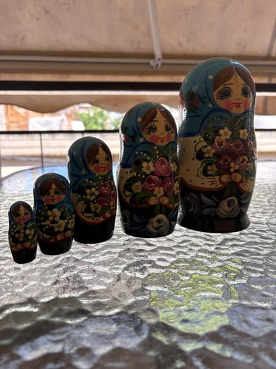 The image shows a set of five Russian nesting dolls placed on a textured glass surface, possibly a table, outdoors. The dolls are arranged in descending order of size from right to left. Each doll is intricately painted with a woman's face wearing a blue headscarf and holding a bouquet of flowers. The background is blurred, showing what appears to be an outdoor setting with some wooden structures and greenery, suggesting it might be a patio or garden area. The lighting indicates it's daytime. The image does not depict any specific location in Ilion, Greece.