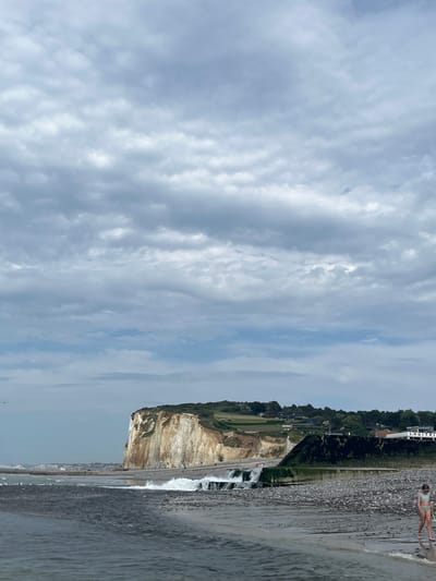 The image shows a view of the beach and cliffs in Hautot-sur-Mer, France. The sky is overcast with dramatic clouds. In the foreground, the dark water of the sea meets a pebble beach. Further inland, a waterfall cascades over a man-made structure. To the left, a sheer white cliff face rises, topped with green fields and trees. In the distance, more coastal cliffs are visible. A small child in a swimsuit is walking on the pebble beach in the right foreground, holding a green stick. Several birds are visible on the water's edge in the mid-ground. Buildings are sparsely located along the coastline.