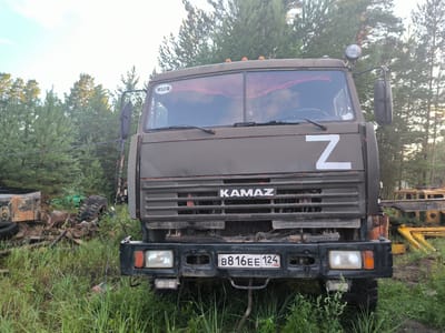 The image shows a dark green KAMAZ truck parked in a grassy, wooded area. The truck has a large grille with "KAMAZ" written on it. A white "Z" symbol is taped to the front of the truck. The truck's windshield has a decorative red curtain with small red balls. A "RUS" sticker is visible on the windshield. The license plate reads "B816EE 124". To the left of the truck, there are other vehicles and debris, including tires and metal parts. The background consists of tall pine trees under a bright sky. The scene appears to be in a rural or industrial area, likely in Russia given the license plate and "RUS" sticker. There are no people visible in the image.