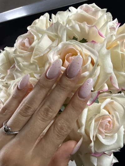 A close-up image captures a person's right hand resting gently on a bouquet of light-colored roses, potentially in Zvezditsa, Bulgaria. The roses are primarily white or very pale cream, with some petals featuring subtle pink or magenta edges and soft pink centers, suggesting a delicate and fresh appearance. The hand displays manicured almond-shaped nails, painted with a light pink polish and accented with small, multi-colored glitter flecks. A silver-toned ring, possibly an engagement or wedding band, with a distinct V-shaped design and small embedded stones, adorns the ring finger. The background is largely dark, which highlights the brightness of the roses and the hand. A vertical light grey or silver bar is partially visible on the left edge of the frame. The image emphasizes details like the nail art, the ring, and the texture of the rose petals, conveying a sense of elegance and celebration, likely associated with a special event. No text is visible in the media file.