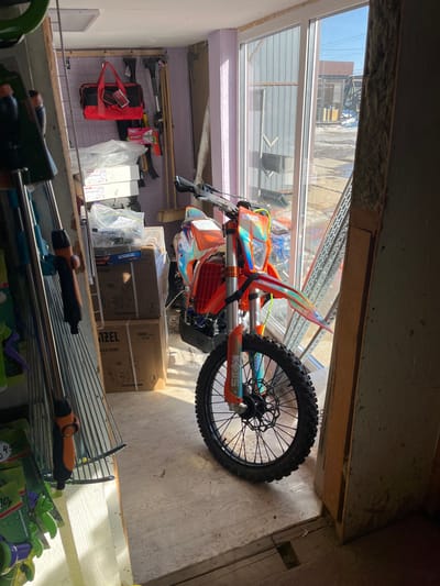 The scene depicts the interior of a structure, identified as being in Popovka, Russia, characterized by a mix of storage and display elements. A prominent orange and iridescent off-road motorcycle, consistent with an enduro or dirt bike, is positioned on a light-colored floor, angled towards a large glass door or window unit. The motorcycle features a black front wheel with a spoked rim and disc brake, orange front fender, forks, and frame components, alongside multi-colored, holographic-effect fairings.
To the left of the motorcycle, a vertical metal shelving unit displays various gardening tools, including rakes with black and orange handles and multiple green spray nozzles. Behind this unit, the back wall, painted a light lavender hue, features a wall-mounted grid panel from which a red fabric tool bag hangs. Several long-handled tools, including a broom, are leaned against the wall. Cardboard boxes, some explicitly labeled "DENZEL WATER PUMP," are stacked on the floor and against the left wall, alongside bundles of white, plastic-wrapped items.
Bright sunlight streams through the glass door/window on the right, casting distinct shadows on the floor and illuminating the motorcycle. Through the clear glass, an exterior environment is visible: patches of white snow or ice are present on the ground, indicating cold weather. Further back, a large building with gray corrugated metal siding and a brown-roofed structure are discernible under a clear blue sky. The immediate right wall within the interior appears unfinished or partially constructed, exposing insulation material. No individuals are present in the image.