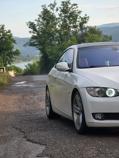 This media file shows the front passenger side of a white car parked on a wet, asphalt road. The car's headlight is on and illuminates the surroundings. Drops of water are visible on the car's body and windshield, indicating that it has recently rained.
Behind the car, lush green trees and a body of water can be seen, with mountains in the distance. The sky appears overcast, suggesting a cloudy or rainy day. The overall scene depicts a quiet, natural setting, possibly a rural area in Rakilovtsi, Bulgaria. There is a small, yellow sign with some unreadable text visible in the background. The lighting suggests it is daytime, but the overcast sky might make it appear dimmer.