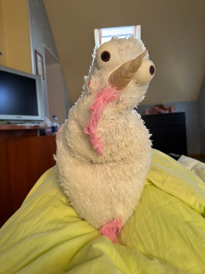 A close-up, eye-level, medium shot captures a white, plush toy with a conical horn and large, round, dark-brown eyes. Pink wisps of material, likely yarn, are attached to the toy's head and body. The toy is positioned on a lime-green surface that appears to be bedding. The background is softly blurred, showing a sloped ceiling, a window with a view of the sky, a dark dresser, and part of a wooden desk with a monitor on it. The lighting suggests an indoor environment, possibly a bedroom. The location of Жостово, Russia, is not visually discernible in the image.