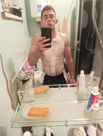 The image shows a man taking a mirror selfie in a bathroom. He is shirtless and wearing black and white shorts, revealing his toned physique. The bathroom appears to be modest, with tiled walls, a toilet, a shower stall, and a sink with a mirror. Various toiletries, including toothpaste and toothbrushes, are visible on shelves and around the sink. A towel is draped over a heating radiator. The man is holding a black smartphone to take the photo. The overall impression is a casual, personal moment captured in a typical bathroom setting. No specific details about the city of Fastiv, Ukraine are visible in the image.