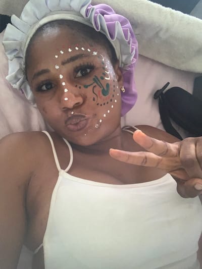 An adult female is depicted in a close-up, upward-angled selfie, presumably taken indoors in Kubi A, Nigeria. She has a dark complexion and wears a ruffled, two-toned satin bonnet, white on the outer layer and lavender on the inner, covering her hair. Her face is adorned with white dot patterns along the bridge of her nose, forehead, cheeks, and chin. On her right cheek, an additional abstract design in green, white, and a darker hue is present. She has extended eyelashes and her lips are pursed. She is wearing a white spaghetti strap tank top. Her left hand is raised, forming a peace sign gesture with her index and middle fingers. The background is indistinct, showing a light-colored fabric behind her and a dark object, possibly a strap or bag, to her right.