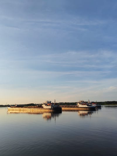 Two cargo barges are on a wide river, reflecting in the calm water under a pale blue sky with thin clouds. The barges are positioned horizontally, one slightly behind the other and to the right. The barge in the foreground, on the left, is loaded with what appears to be cargo, and its hull is illuminated by the warm light of the setting sun, creating a golden reflection on the water. The second barge, to the right and slightly further back, has a superstructure with cabins and lifeboats visible. The distant shore is lined with a dark green band of trees. The scene suggests a serene evening on a waterway in Shchetinino, Russia.