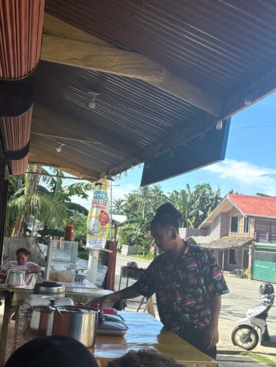 This image depicts an outdoor or semi-enclosed eatery located in Burgos, Philippines, under bright daylight conditions.

A person with dark dreadlocks, wearing a short-sleeved patterned shirt and black ear gauges, stands behind a counter, extending their right hand towards a lidded container on the counter. Visible on the counter are multiple lidded stainless steel pots, additional lidded rectangular containers, a stack of light-colored cups, and a clear glass display case with unidentified contents. To the left, a second individual, a woman in a light-colored t-shirt, is seated at a table, looking downwards. The eatery's structure includes a corrugated metal roof, exposed wooden beams, and multiple visible lightbulbs.

Beyond the eatery, an asphalt road curves to the right, bordered by dense tropical vegetation, including numerous palm and banana trees. A tall, colorful banner advertising "WOMERS PANAO SALE!" is erected roadside on the left. On the right side of the road, a two-story house with a red-orange corrugated roof and brick-patterned siding is visible, positioned above a smaller structure with a textured brown roof. A white scooter with a helmet on its seat is parked on the roadside near the houses. The sky is clear blue with minimal cloud cover, and overhead utility lines are discernable.