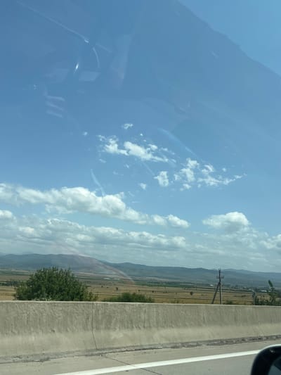 A view from a vehicle on a sunny day shows a rural landscape in Kvemo Akhalsopeli, Georgia. In the foreground, a concrete highway barrier runs horizontally across the frame. Beyond the barrier, a field of dry, golden crops stretches towards rolling hills in the distance. A single green tree stands prominently in the middle ground. A power line pylon with multiple wires is visible to the right of the tree. The sky is a clear blue with scattered white cumulus clouds. The reflections of the vehicle's interior, including what appears to be a steering wheel, are visible on the windshield. The overall scene depicts a bright, outdoor environment with agricultural land and a clear sky.