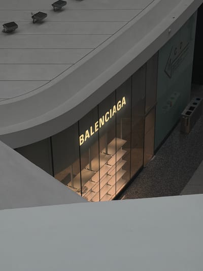 The image shows an exterior view of a Balenciaga store. The store has a modern design with a large glass facade. The Balenciaga logo is prominently displayed in illuminated letters on the facade. Inside the store, empty shelves are visible. The store is situated in a public area with a grey, speckled pathway leading up to it. Above the store, a curved architectural element can be seen. In the background on the right, a trash receptacle with multiple compartments is present. The upper portion of the image shows a grey, tiled surface with several spotlights. There are no people visible in the image. The specific location in Guangyang District, China, is not discernible from the visual information provided.