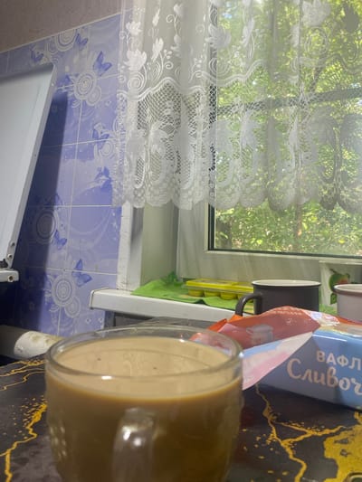 The image depicts a close-up view of a glass mug filled with coffee, placed on a dark countertop with a gold marble-like pattern. In the background, a window is visible with a white lace curtain, through which lush green foliage can be seen, suggesting an outdoor scene with trees. On the windowsill, there is a yellow ice cube tray, a dark gray mug, and another mug with a picture of an apple. To the left of the window, a tiled wall with a blue and white butterfly pattern is visible. The overall impression is a domestic setting, possibly a kitchen, with a beverage ready to be consumed. The location is identified as Shuya, Russia.