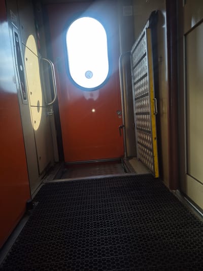 The image shows the interior of a train carriage, specifically the area near a door. A large, oval window with bright light shining through it dominates the center of the frame. To the left of the window is a closed, light-colored door with a silver handle and several circular indentations. To the right of the window is an open door with a yellow-trimmed metal panel, revealing a section of the train's interior. The floor is covered with a black, honeycomb-patterned mat. The overall impression is that of a utilitarian interior space within a mode of transport. There is no discernible information within the image to suggest a location in Turkistan, Kazakhstan.