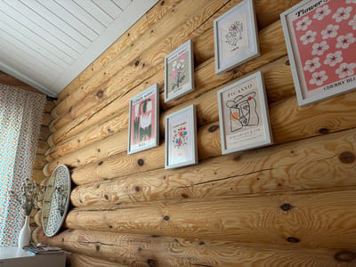 In a rustic room with log walls, several framed prints are displayed. One print features a line drawing of a dancing figure with a flower for a head, attributed to K. Haring. Another print depicts pink cherry blossoms on a pink background, with the text "CHERRY BLO" visible. To the left, a print shows a stylized floral arrangement with the text "FLOWER MARKET" and "ROME." Below this, another print displays abstract floral shapes in pink and green, with "IBIZA" written at the bottom. Adjacent to the "IBIZA" print, a smaller print features a colorful illustration of flowers and the text "OSLO." Further to the right, a print shows a cubist-style portrait with the text "PICASSO" and information about an exhibition at Berggruen & Cie in Paris from February to March 1953. On the left side of the image, a white vase holds dried flowers, and a round mirror hangs on the log wall. A patterned curtain is visible behind the vase. The ceiling is made of white wooden planks. The scene is located in Shashmurka, Russia.