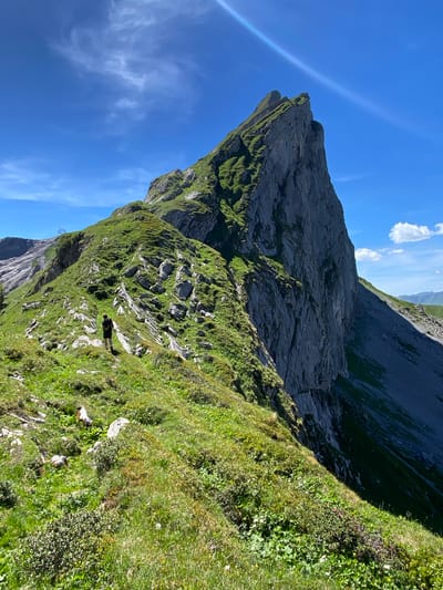 A lone hiker is walking on a grassy mountain trail in the Alps. The hiker is wearing a dark t-shirt and shorts and carrying a backpack. The trail winds along a steep, green mountainside with rocky outcrops. In the background, a massive, jagged rock face dominates the landscape, bathed in bright sunlight under a clear blue sky with wispy clouds. The scene is in a mountainous region, consistent with the area around Brienz, Switzerland.
