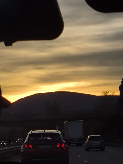 The image captures a sunset scene from inside a vehicle on a road in Niedernai, France. The upper portion of the frame is obscured by dark, curvilinear vehicle interior components, possibly a dashboard or rearview mirror housing. The sky displays a gradient of golden-orange and light gray hues, with visible horizontal cloud streaks. Directly in front of the observer's vehicle is a red SUV, identifiable by its illuminated red taillights and a French license plate reading "SG-093-LO". To the right of the red SUV, in an adjacent lane, a white box truck is visible. Further ahead in the right lane, a dark-colored sedan or hatchback is present. Distant vehicle headlights are faintly visible in the far left lane. The road surface is asphalt with white lane markings. In the background, dark, silhouetted hills or mountains are visible against the bright horizon line, with sparse trees appearing on the slopes and to the far right. No people or specific interactions are discernibly depicted. The environment suggests an open road or highway setting at dusk.