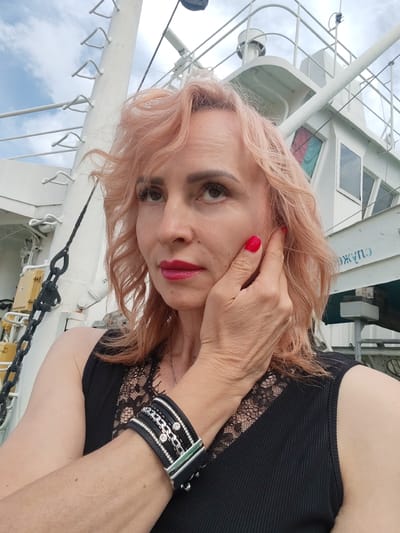 The photograph is a close-up, waist-up, selfie-style portrait of a Caucasian woman with light pink, wavy hair. She has fair skin, dark eyebrows, and is wearing dark eyeliner and bright pink lipstick. Her right hand is touching her cheek, and her fingernails are painted red. She is wearing a black tank top with lace detailing on the neckline and a black bracelet with silver accents and clear stones on her left wrist.
The background appears to be a large white vessel, possibly a ferry or ship, with a metal railing and a chain anchor visible on the left. The sky is overcast with some blue patches visible. The text "ЖАУПО" is visible in blue lettering on the side of the vessel. The location is identified as Faliukovo, Russia.