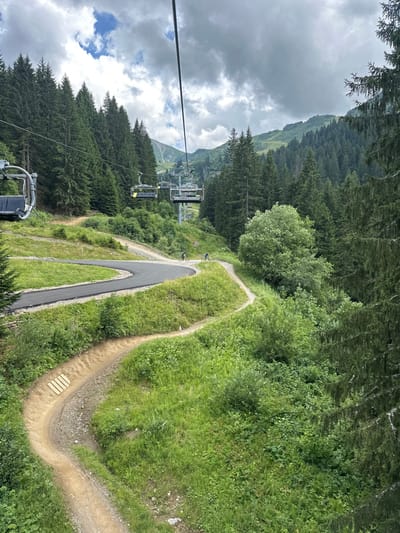 An aerial view from a ski lift chair depicts a summer mountain landscape in Châtel, France, under an overcast sky. The foreground features a narrow, winding dirt mountain bike trail with wooden steps embedded in its surface, alongside a wider, dark asphalt-paved switchback trail. Lush green grass and varied green foliage cover the slopes. In the mid-ground, two individuals are visible on mountain bikes, riding downhill on a dirt trail that parallels the paved section. A large grey ski lift support pylon stands centrally, with multiple empty ski lift chairs suspended from its cables, extending into the background. Dense, dark green coniferous forests dominate the mid-ground and background mountainsides, with higher peaks visible in the distance. The scene captures active summer recreational use of the mountain, facilitated by the ski lift and designated bike trails.