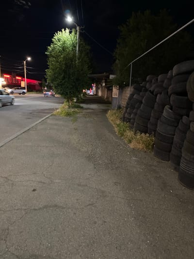 The image depicts a night scene on a street in the vicinity of the Border Gyumri-Akhuryan in Armenia. A sidewalk runs alongside the street, which appears to be paved with asphalt and shows signs of wear. To the right of the sidewalk, a large pile of old tires is stacked against a low wall. Beyond the wall, trees are visible. On the left side of the street, cars are parked and driving. Buildings with illuminated signs, appearing to be businesses, are also present on that side of the street. A streetlamp illuminates the scene, casting a bright light on a nearby tree. The sky is dark, indicating nighttime. There are no people clearly visible in the image, but their presence is implied by the cars and the urban setting.