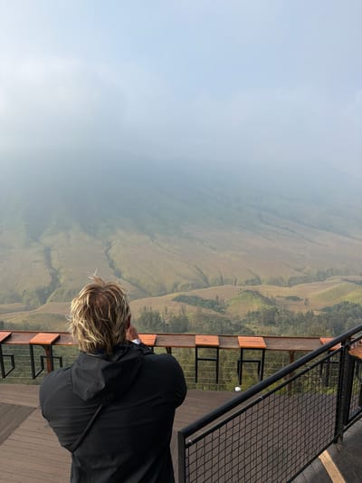 A person with blond, wavy hair stands with their back to the camera, wearing a black hooded jacket. They are on a wooden deck, facing a vast, hilly landscape partially obscured by fog. In front of them is a railing and a series of wooden tables attached to a railing. The hills in the background are covered in dry, golden grass with some green patches and trees. The sky is overcast with heavy fog. This scene appears to be from a scenic viewpoint in Ngadas, Indonesia.