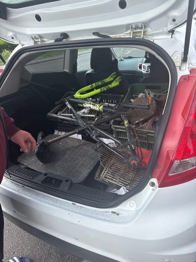 A medium shot shows the open trunk (boot) of a white car, likely in Boston, United Kingdom, filled with various items. A person's hand, wearing a dark red long-sleeved shirt, is visible in the foreground, reaching into the trunk, suggesting loading or unloading activity.

The trunk is packed with a partially disassembled or well-used black bicycle frame featuring a prominent yellow-green front fork. A rusty chain is visible on the bicycle, and the word "DEFEKTR" is partially visible on the bike's frame. Several green plastic crates or baskets are stacked around the bicycle, along with a metal wire basket, possibly from a shopping trolley. A black, ribbed rubber car mat lies on the trunk floor.

The car's exterior is white, with a red rear light cluster visible at the top of the image. The rear bumper, equipped with at least two visible parking sensors, is seen at the bottom right. Minor debris, possibly dry grass, is scattered along the trunk's rim. Through the open trunk, a glimpse of the car's interior, including the rear seats and a door panel, is visible. The scene is set outdoors during daylight hours, with an asphalt surface visible near the car, indicating a parking area or street. The items in the trunk suggest the transportation of goods, possibly salvaged or used materials.