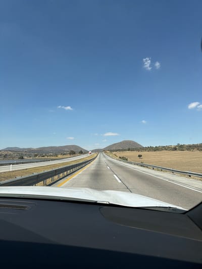 A view from inside a vehicle looking down a long, straight highway under a bright blue sky with scattered clouds. The road is divided by white lines, and guardrails run along the sides. The landscape features dry, grassy fields and rolling hills in the distance. The interior of the car is visible in the foreground.