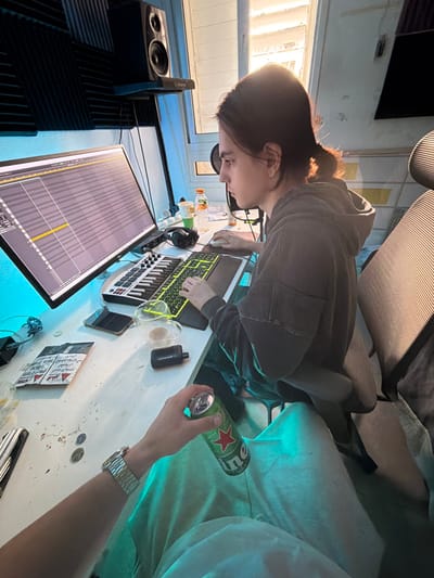 The image depicts an indoor scene in Migdal HaEmeq, Israel, appearing to be a home studio or workspace. In the mid-ground, a person with dark hair tied back, wearing a dark hooded sweatshirt, is seated in a gray office chair, intently focused on a large computer monitor displaying a digital audio workstation interface with a visible musical timeline. Their right hand operates a computer mouse, and their left hand rests on a green-backlit keyboard. A small MIDI keyboard controller is positioned in front of the monitor.
To the left of the monitor, an Alesis studio monitor speaker is mounted on a stand, and blue LED strip lighting illuminates the acoustic foam panels on the wall. To the right of the monitor, a microphone with a pop filter is visible. The desk surface, primarily white, holds various items: a dark smartphone, a glass mug containing a light brown liquid, a dark vape device, a plastic bottle of orange beverage, several Israeli shekel coins, and a white packet with red Arabic text.
In the foreground, a partial view of another individual is present, showing their right arm, wearing a silver-toned wristwatch, and their lap, dressed in light-colored fabric. This individual holds a green Heineken beer can. Natural light streams in from a window in the background, illuminating the scene and creating shadows from blinds on the wall.