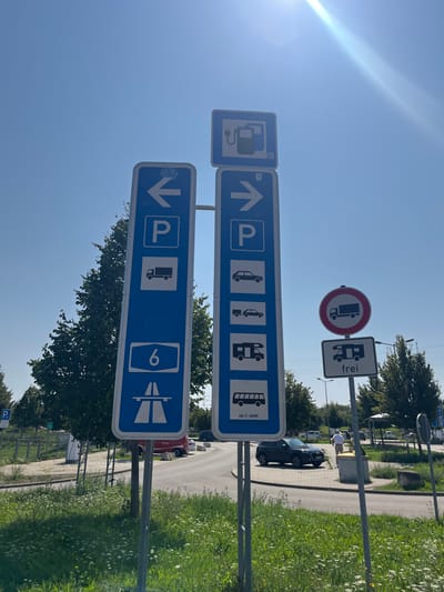 The scene depicts a sunny outdoor environment at a roadside service or rest area, specifically in the vicinity of Sinsheim, Germany, indicated by signage for Autobahn 6.

In the foreground, a patch of green grass is visible, interspersed with small white flowers. Dominating the center are two tall, vertical blue-and-white German road signs mounted on metal poles. The left sign features a left arrow, a "P" for parking, a truck symbol, and a blue Autobahn shield with the number "6". The right sign displays a right arrow, a "P" for parking, a car symbol, a car with trailer symbol, a recreational vehicle (RV) symbol, and a bus symbol, with "130 ET JAHRE" inscribed below it. Above the right sign, a smaller blue-and-white square sign indicates an electric vehicle charging station. To the right of these main signs, a circular red-and-white "no trucks" prohibition sign is mounted above a rectangular black-and-white sign displaying an RV symbol and the word "frei" (allowed/free).

In the mid-ground, a paved asphalt road curves, with a dark-colored SUV (likely an Audi) parked on the right side. Another red vehicle is partially visible further left. One adult male, dressed in a light-colored shirt and dark trousers, is seen walking away from the camera on a paved path near a gray metal waste receptacle or bollard. The background features various deciduous trees with green foliage and additional paved areas, suggesting a larger parking or service facility. The sky is clear blue with a prominent sun flare in the upper right quadrant, indicating bright daylight.