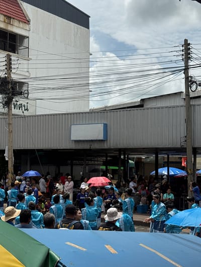 The image depicts an outdoor scene in Prakhon Chai District, Thailand, possibly during a festival or parade. A crowd of people, many wearing bright blue traditional outfits, are gathered. Some individuals hold umbrellas, one blue and two pink, providing shade. The background features a building with a red-tiled roof and white walls, along with a silver corrugated metal structure. A large cluster of electrical wires hangs prominently in the foreground, crisscrossing the scene. A sign with green lettering is visible on the white wall of the building. The overall impression is one of a lively community event.