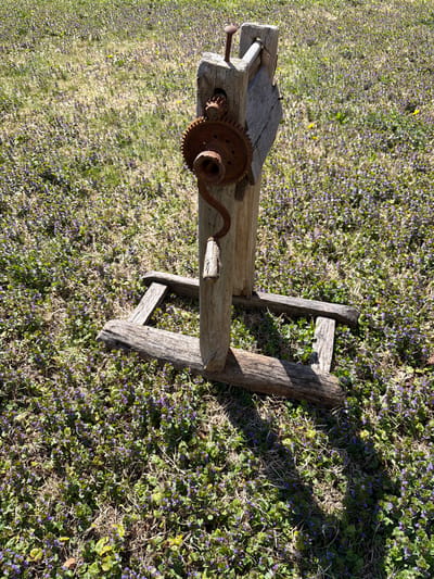 An old, wooden, possibly handmade, piece of farm machinery sits on a field of ground cover with small purple flowers. The device has a handle, gears, and appears rusty. The location is unspecified but it's supposed to be in the city of Valchedram, Bulgaria. The setting is outdoors, likely during the day based on the lighting. No people are visible.