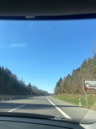 The image shows a view from a car driving on a highway. The sky is a clear blue, and the highway is bordered by trees on both sides. There is a sign on the right side of the road that reads "Villa Rustica Mehring". The road is straight and the environment is rural. The image was taken in Mehring, Germany, at the coordinates 49.763, 6.859. The car is driving towards a bridge in the distance. The car is in the right lane of the highway. The highway is four lanes wide, with two lanes in each direction. There is a white line marking the separation of the two lanes. The road is paved with asphalt, and there are white lines painted on the surface. The trees are deciduous and are bare of leaves, suggesting that it is autumn or winter. The trees on the left side of the road are closer to the car than the trees on the right side. The sun is shining, and the shadows of the trees are visible on the road.
