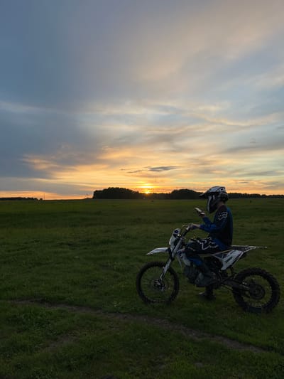 A medium, outdoor shot depicts a man on a dirt bike against a colorful sunset. The location is an open, grassy field. The man, wearing a helmet and riding gear, is partially visible and appears to be interacting with the bike. The dirt bike is a prominent object. The sunset is a key environmental detail, coloring the sky with shades of orange and grey. The ground shows signs of tire tracks in the grass. The location cannot be determined to be Новочувашский, Russia.